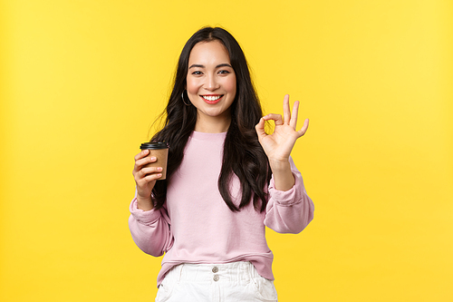 People emotions, lifestyle leisure and beauty concept. Smiling good-looking asian girl with takeaway cup show okay gesture, recommend great cafe with delicious coffee and desserts, yellow background.