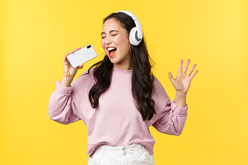 People emotions, lifestyle leisure and beauty concept. Carefree happy asian woman in headphones, playing karaoke app on mobile phone, using smartphone as microphone and singing along song.