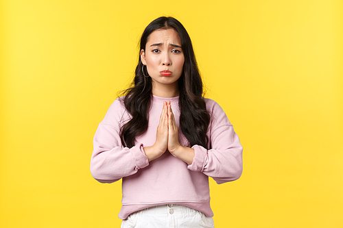 Lifestyle, emotions and advertisement concept. Pitiful cute asian girl pouting and hold hands in plead, begging for favor or help, asking for something, feeling sorry, standing yellow background.