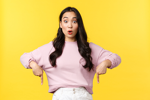 People emotions, lifestyle and fashion concept. Astonished and impressed pretty asian woman in stylish outfit pointing fingers down to show cool promo offer, advertise product over yellow background.