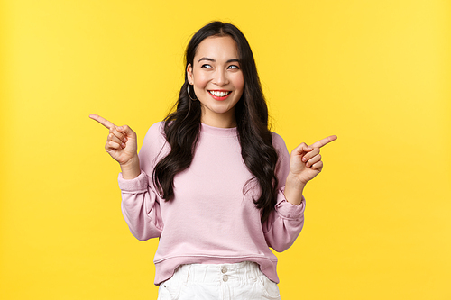 People emotions, lifestyle, fashion concept. Excited smiling pretty asian woman with happy expression, looking left while pointing sideways and promote two variants, both products, yellow background.