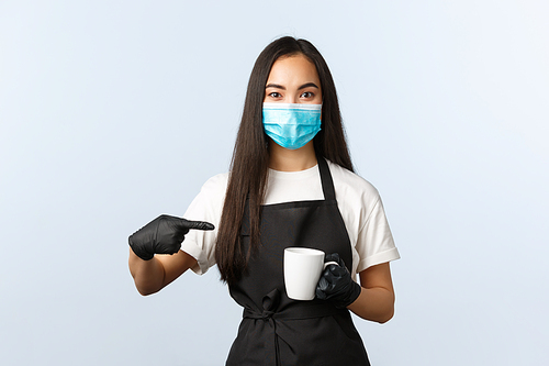 Covid-19, social distancing, small coffee shop business and preventing virus concept. Smiling asian barista in medical mask and gloves recommend try coffee at her cafe, pointing at cup.
