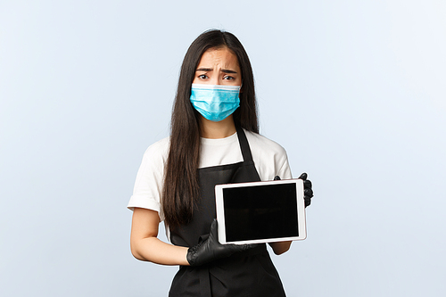 Covid-19 pandemic, social distancing, small business and preventing virus concept. Upset gloomy asian waitress, cafe staff in medical mask and gloves frowning sad and showing digital tablet display.