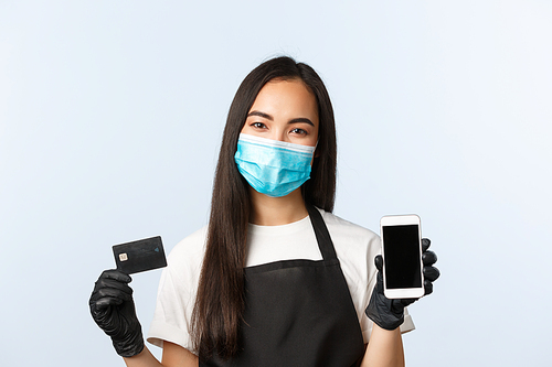 Covid-19 pandemic, coffee shop, small business and preventing virus concept. Smiling pleasant asian female worker, cafe barista in medical mask show smartphone and credit card.