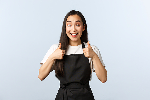 Coffee shop, small business and startup concept. Excited smiling asian waitress approve new menu. Cute female barista in black apron show thumbs-up in support, like or agree.