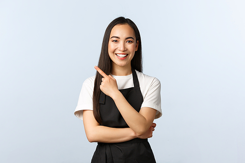 Coffee shop, small business and startup concept. Cheerful asian female working part-time at cafe, wear black apron, pointing finger upper left corner, inviting see banner or advertisement.