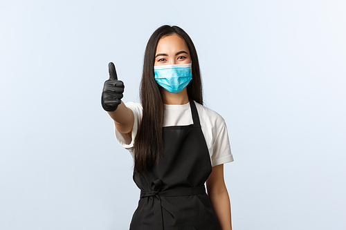 Covid-19, social distancing, small coffee shop business and preventing virus concept. Smiling asian female barista, employee in medical mask and gloves show thumb-up.