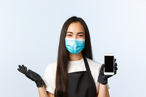 Covid-19 pandemic, coffee shop, small business and preventing virus concept. Smiling cute asian female barista showing cafe application on smartphone screen, bonuses and special discount promo.