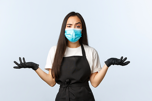 Covid-19, social distancing, small coffee shop business and preventing virus concept. Confused and annoyed asian employee, barista in medical mask and gloves shrugging with spread hands sideways.