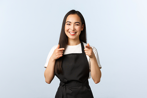 Coffee shop, small business and startup concept. Cheerful smiling female barista, asian cafe staff in black apron pointing at camera, inviting consumers visit and try new desserts, white background.