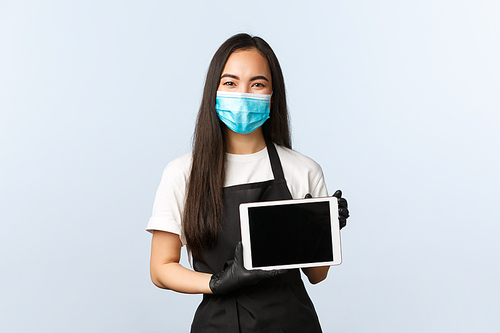 Covid-19 pandemic, social distancing, small business and preventing virus concept. Friendly smiling female asian barista in medical mask and gloves, showing digital tablet, how to make order online.