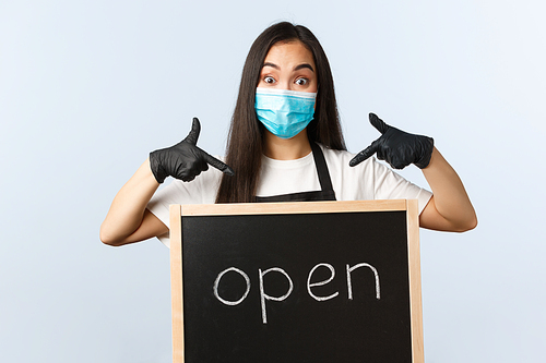 Small business, covid-19 pandemic, preventing virus and employees concept. Excited cute asian female barista, cafe or store worker in medical mask pointing at we are open sign.
