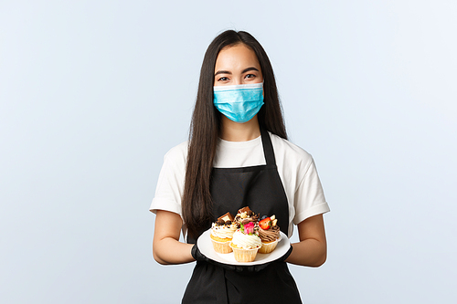 Covid-19, social distancing, small coffee shop business and preventing virus concept. Smiling cute asian cafe owner, female barista or waitress bring order with muffins on plate, wear medical mask.