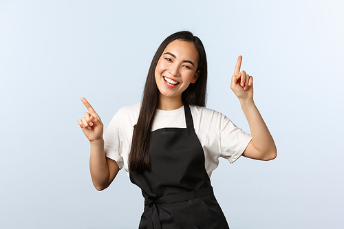 Coffee shop, small business and startup concept. Rejoicing attractive female asian employee in black apron dancing and having fun, singing, pointing fingers up, white background.