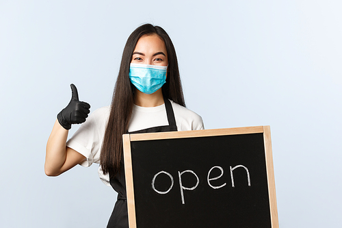 Small business, covid-19 pandemic, preventing virus and employees concept. Smiling cute asian female employee, cafe or store worker holding open sign, invite visitors.