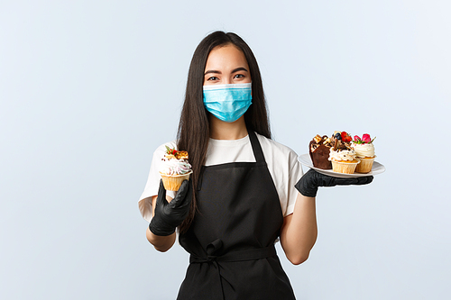 Covid-19, social distancing, small coffee shop business and preventing virus concept. Friendly cute asian barista, worker in medical mask handing you muffin, serving order in cafe.