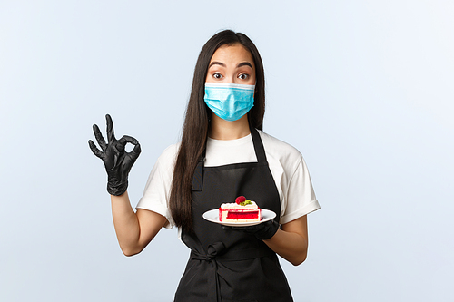 Covid-19, social distancing, small coffee shop business and preventing virus concept. Cute asian waitress, female barista in medical mask and gloves, show okay sign and give dessert cake.