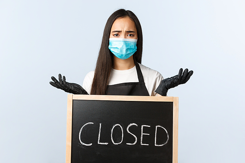 Small business, covid-19 pandemic, preventing virus and employees concept. Complicated and confused asian employee, barista in medical mask shrugging over closed sign.