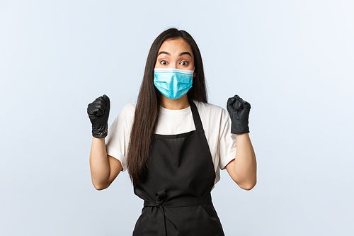 Covid-19, social distancing, small coffee shop business and preventing virus concept. Cheerful happy asian employee, female barista clench fists in rejoice, wear medical mask and gloves.