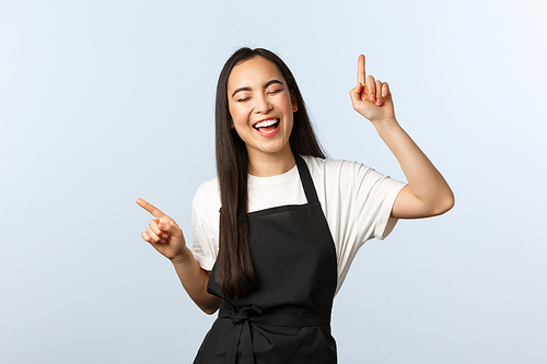 Coffee shop, small business and startup concept. Carefree cheerful asian female barista, grocery store employee in black apron, singing adn dancing with fingers pointing top, have fun happy.