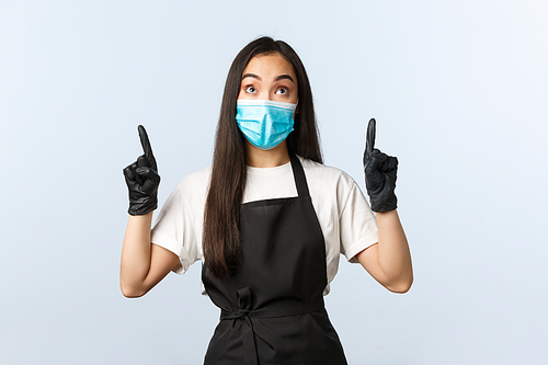 Covid-19 pandemic, social distancing, small business and preventing virus concept. Excited asian female barista in medical mask and gloves seeing cool ad, pointing fingers up.