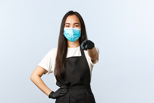 Covid-19, social distancing, small coffee shop business and preventing virus concept. Cheerful asian employee, restaurant barista in medical mask and gloves pointing at camera, invite consumers.
