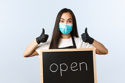 Small business, covid-19 pandemic, preventing virus and employees concept. Upbeat asian female store worker, cafe staff or barista in medical mask show thumb-up and we are open sign.