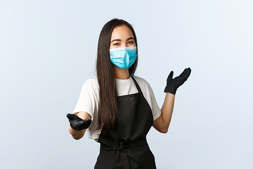 Covid-19, social distancing, small coffee shop and preventing virus concept. Charismatic smiling asian employee, waitress or barista inviting visit cafe, pointing at camera and behind, wear mask.