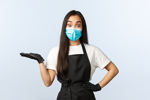 Covid-19, social distancing, small coffee shop business and preventing virus concept. Surprised asian female cafe staff, barista in medical mask and gloves introduce new product on her hand.