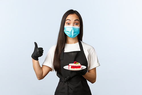 Covid-19, social distancing, small coffee shop business and preventing virus concept. Excited cafe owner, employee in medical mask and gloves, serving delicious cake, show thumb-up.
