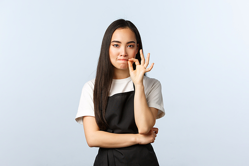 Coffee shop, small business and startup concept. Serious cute korean barista, female employee at cafe promise keep secret, zip or lock lips, standing in black apron.