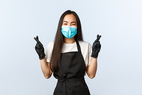 Covid-19, social distancing, small coffee shop business and preventing virus concept. Hopeful and optimistic smiling asian waitress, barista in medical mask and gloves cross fingers good luck.
