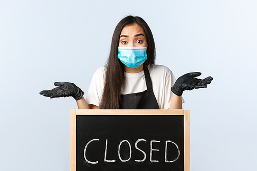 Small business, covid-19 pandemic, preventing virus and employees concept. Confused and puzzled asian female employee in medical mask shrugging, dont know when cafe opens after lockdown.
