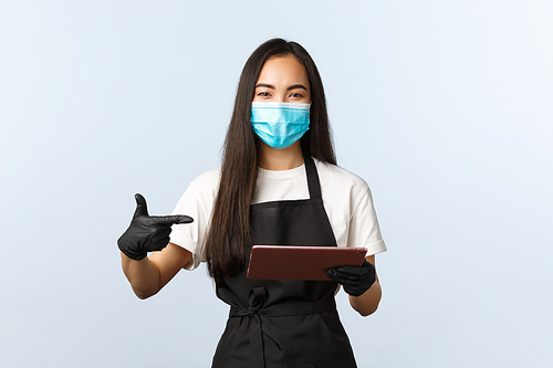 Covid-19, online orders, small coffee shop business and preventing virus concept. Smiling asian barista, waitress in medical mask and gloves pointing at digital tablet, taking consumer order.