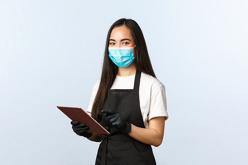 Covid-19, online orders, small coffee shop business and preventing virus concept. Smiling nice asian female barista in medical mask and gloves during coronavirus, listen customer, hold digital tablet.