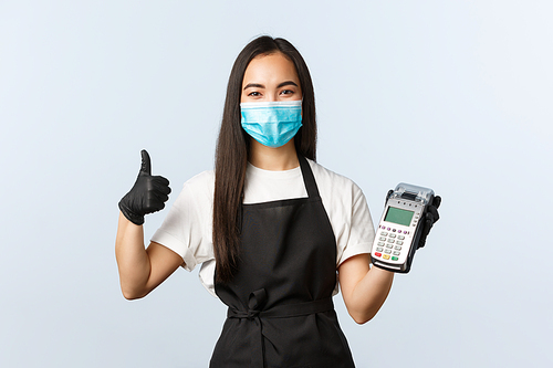 Small business, coronavirus, social distancing and contactless payment concept. Asian barista, waitress in cafe, medical mask and gloves, recommend use POS terminal for safe purchase during covid-19.