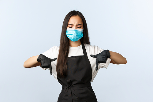 Covid-19 pandemic, social distancing, small business and preventing virus concept. Skeptical and doubtful asian barista, female coffee shop employee in medical mask and gloves pointing down upset.