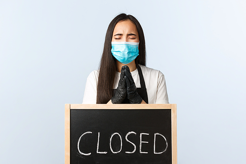 Small business, covid-19 pandemic, preventing virus and employees concept. Hopeful desperate barista, cafe owner in medical mask praying for cafe to open near closed sign during corona lockdown.
