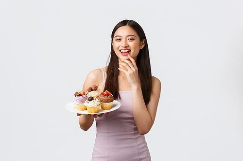 Lifestyle, holidays, celebration and food concept. Beautiful young asian woman host of party in evening dress, treating guests with delicious desserts, holding plate with cupcakes, white background.