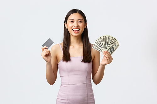 Luxury women, party and holidays concept. Cheerful elegant asian woman in dress, holding money and credit card with happy satisfied face, shopping over white background.