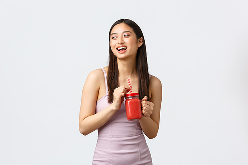 Healthy lifestyle, people emotions and leisure concept. Attractive stylish asian woman enjoying fresh smoothie, wear trendy dress, laughing and looking away dreamy, white background.