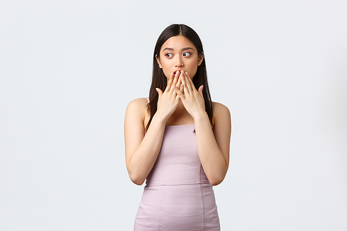 Celebration, beauty and glamour concept. Shocked asian girl in luxurious evening dress, gasping, cover mouth amazed and looking left as gossiping at party about host, white background.