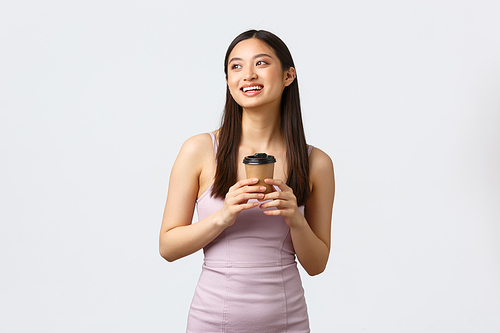 Lifestyle, people emotions and glamour concept. Dreamy asian girl in dress, smiling and looking upper left corner imaging, enjoying beautiful day with cup of coffee, drinking from takeaway cup.