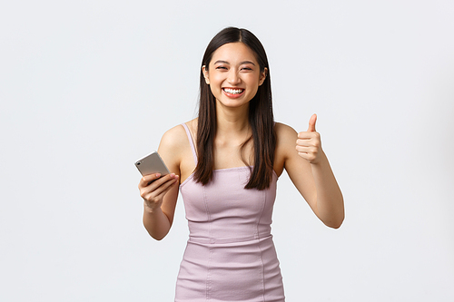 Luxury women, party and holidays concept. Carefree happy asian girl in evening dress, holding mobile phone showing thumbs-up and laughing out loud from funny message or joke, white background.