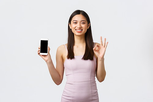 Luxury women, party and holidays concept. Smiling good-looking asian woman in evening dress, recommending application, showing okay gesture and mobile phone screen, white background.