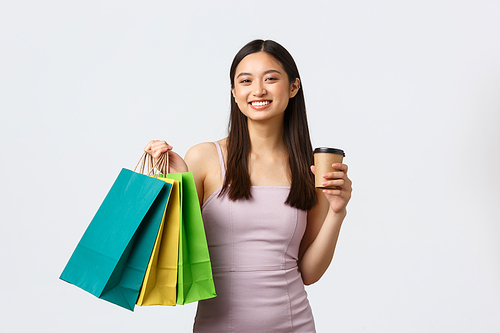 Lifestyle, shopping and tourism concept. Elegant good-looking korean woman in dress, bragging with new clothes, enjoying weekend in mall, drinking coffee, holding bags, white background.