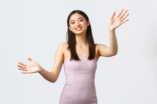 Celebration, beauty and glamour concept. Charming and beautiful asian female host of party in luxurious evening dress, greeting guests, reaching hands for hug and smiling, white background.