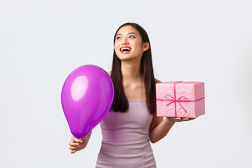 Celebration, party and holidays concept. Dreamy happy birthday girl in evening dress, enjoying b-day and laughing at upper left corner banner, holding gift and balloons, white background.