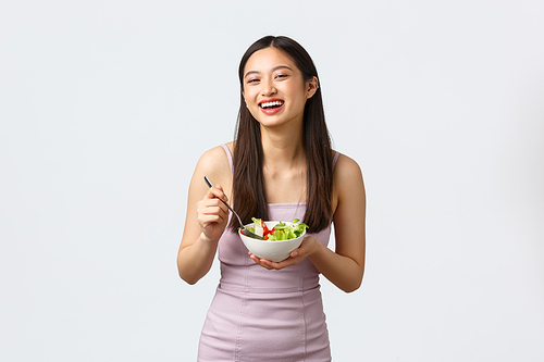 Healthy lifestyle, leisure and people emotions concept. Cheerful smiling asian girl eating salad and laughing happy, wearing party dress, standing white background.