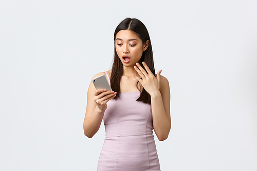 Luxury women, party and holidays concept. Shocked and startled asian girl in evening dress, drop jaw and stare at smartphone screen, reading astounding news in mobile phone app with awe.